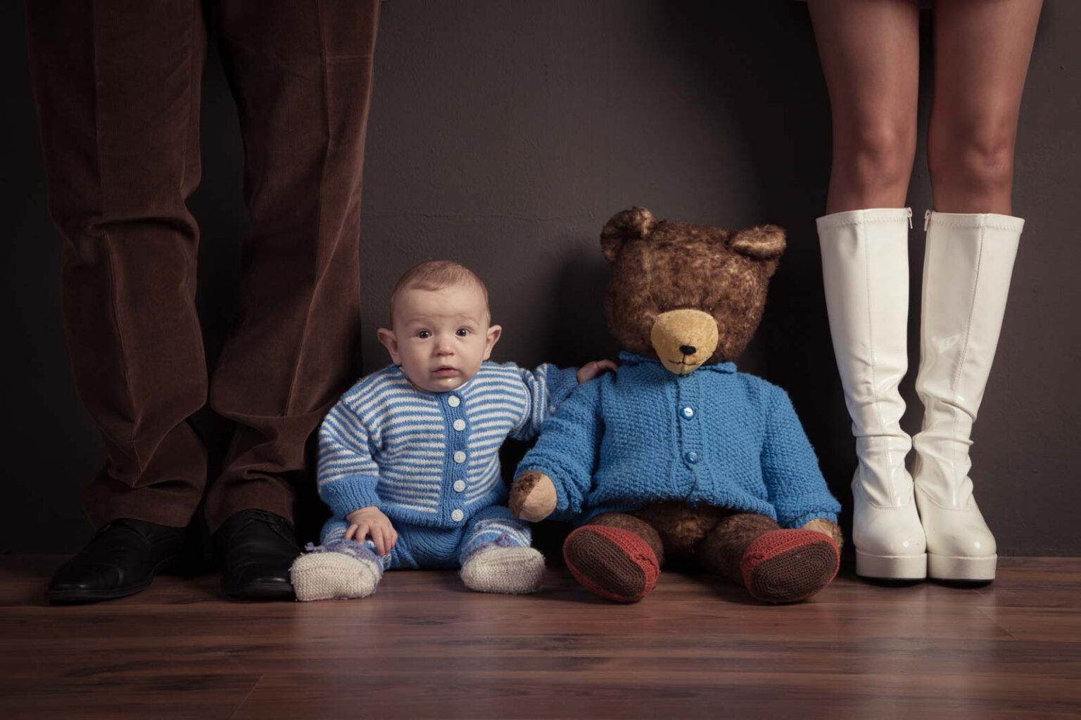 Kleines Baby und großer Teddybär in blauen Strickjacken, flankiert von Erwachsenenbeinen in weißen Stiefeln und braunen Hosen