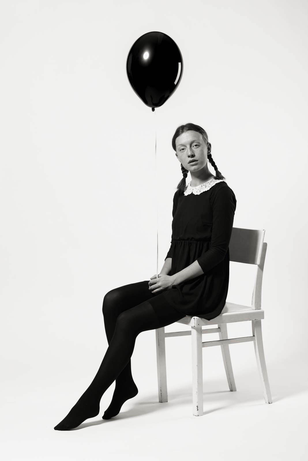 Mädchen mit Zöpfen sitzt auf einem Stuhl und hält einen schwarzen Ballon, gekleidet in ein schwarzes Kleid mit weißem Kragen