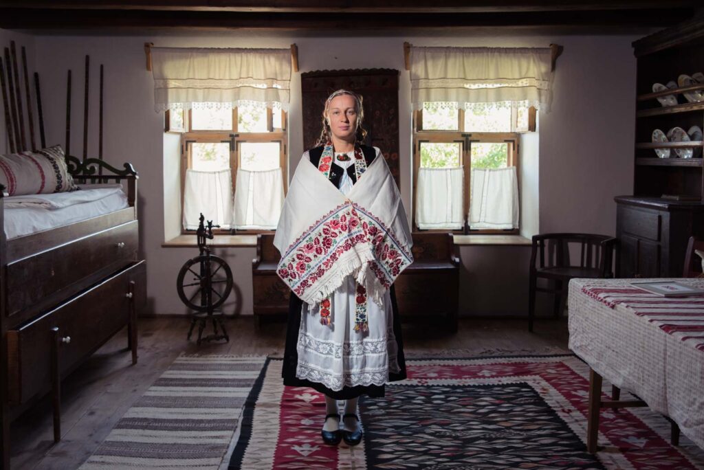 Frau in traditioneller Kleidung steht in einem rustikalen Raum mit Holzmöbeln und einem Spinnrad im Hintergrund