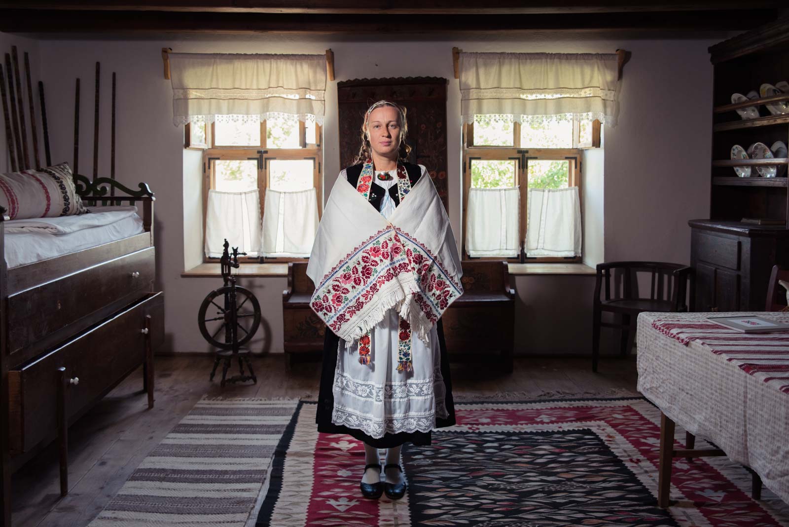Frau in traditioneller Kleidung steht in einem rustikalen Raum mit Holzmöbeln und einem Spinnrad im Hintergrund