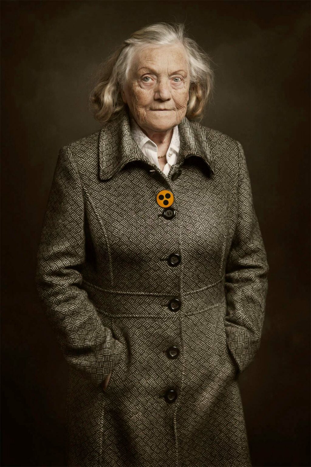 Ältere Frau mit grauem Haar trägt einen gemusterten Mantel und steht mit den Händen in den Taschen vor einem dunkelbraunen Hintergrund; sie trägt ein Emoji-Abzeichen auf ihrem Mantel.