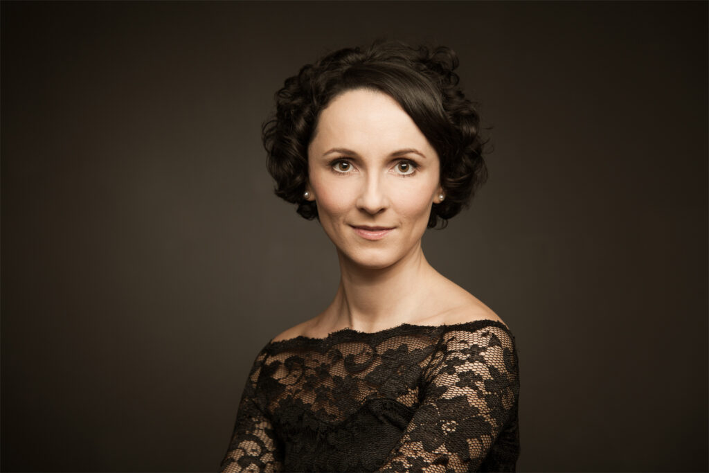 Ein Porträt einer Frau mit kurzem, lockigem Haar, die ein schwarzes Spitzenkleid trägt, vor einem dunklen Hintergrund.
