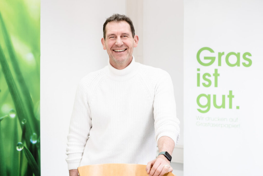 Ein lächelnder Mann in einem weißen Pullover steht an einem Tisch. Im Hintergrund sind zwei Banner zu sehen, eines zeigt Gras mit Tau, das andere trägt den Text 'Gras ist gut. Wir drucken auf Grasfaserpapier!' in grüner Schrift.