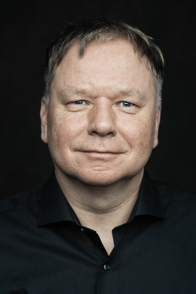Porträt eines Mannes in dunklem Hemd vor dunklem Hintergrund, der direkt in die Kamera schaut.