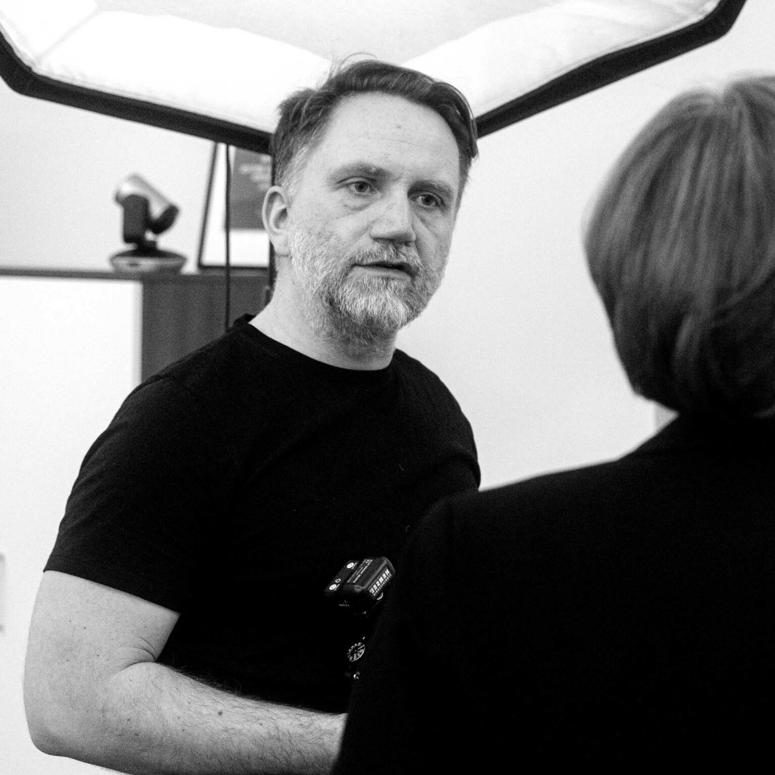 Ein Mann mit Bart in einem schwarzen T-Shirt steht in einem Raum mit Beleuchtung, während er eine andere Person ansieht, die mit dem Rücken zur Kamera steht.