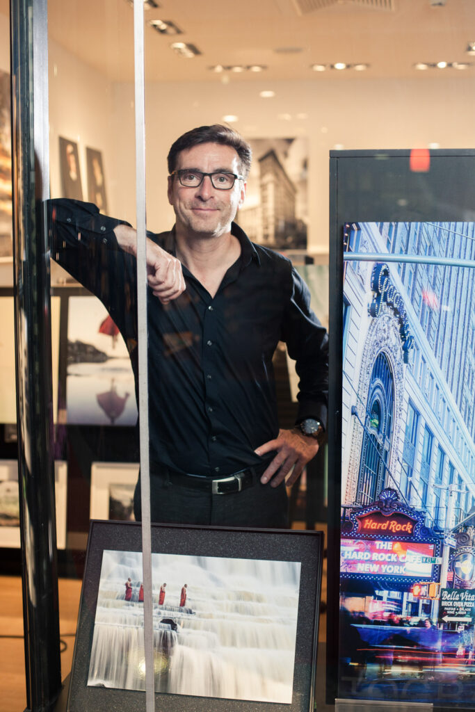 Ein Mann in einem schwarzen Hemd steht in einer Galerie neben Fotokunstwerken, darunter ein Bild mit einem Wasserfall und eines von einem Gebäude in New York.