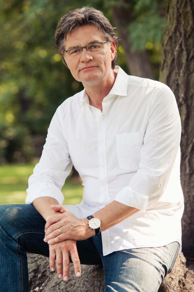 Ein Mann mit Brille und weißem Hemd sitzt im Freien auf einem Baumstumpf, umgeben von grüner Natur.