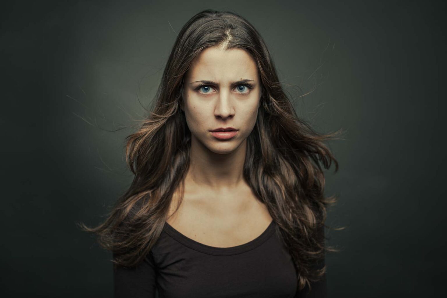Junge Frau mit langen braunen Haaren und ernstem Gesichtsausdruck, trägt ein schwarzes Oberteil, vor einem grauen Hintergrund.