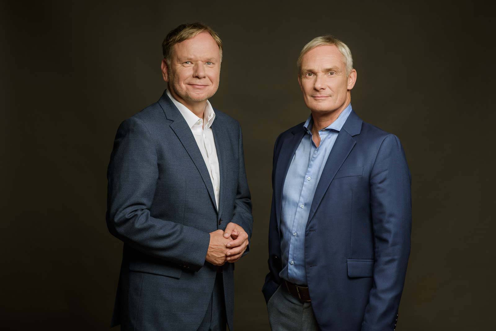 Zwei Männer in Anzügen stehen nebeneinander vor einem neutralen Hintergrund