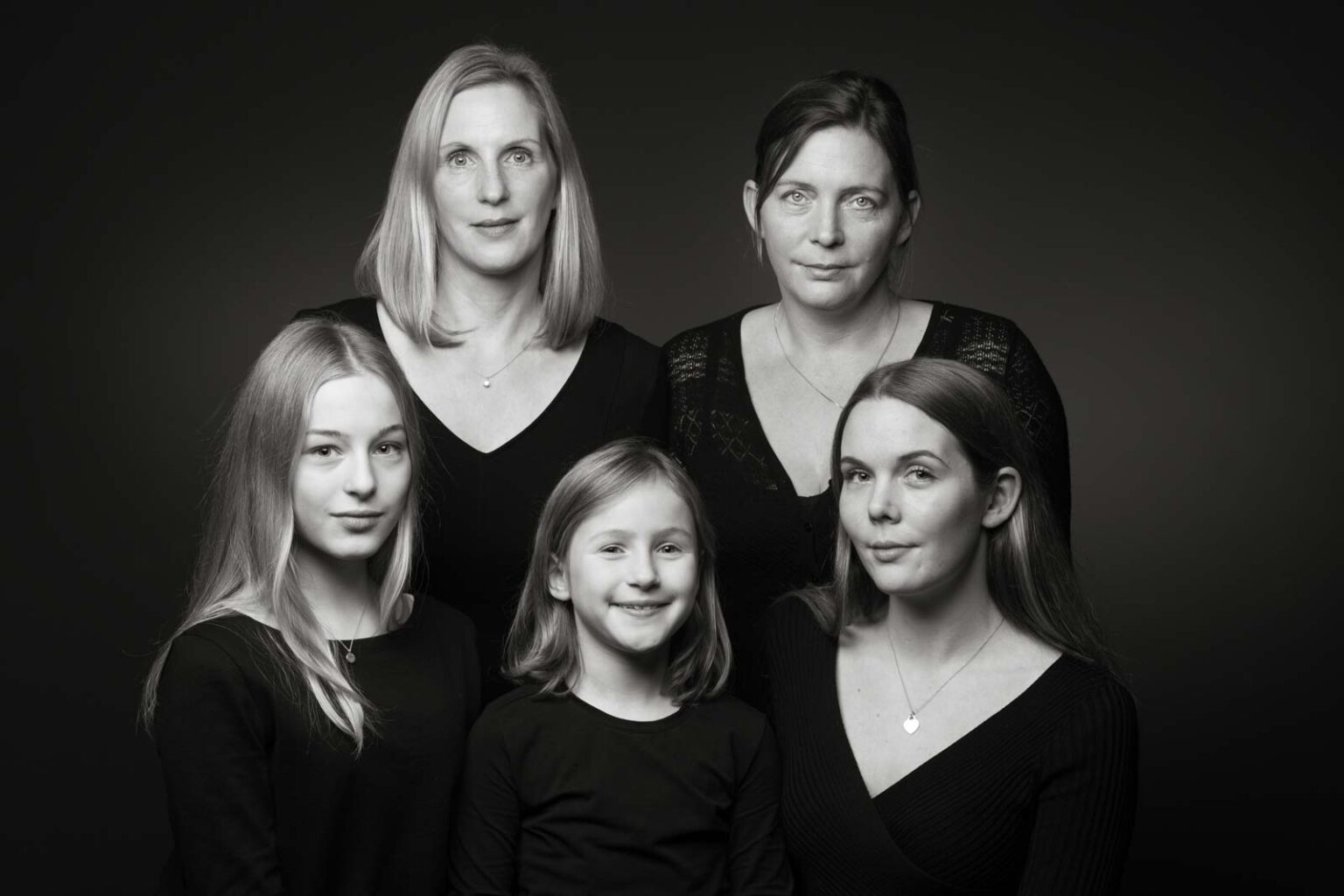 Fünf Frauen unterschiedlichen Alters, alle in schwarzer Kleidung, posieren in einem Studio für ein Schwarz-Weiß-Porträt.