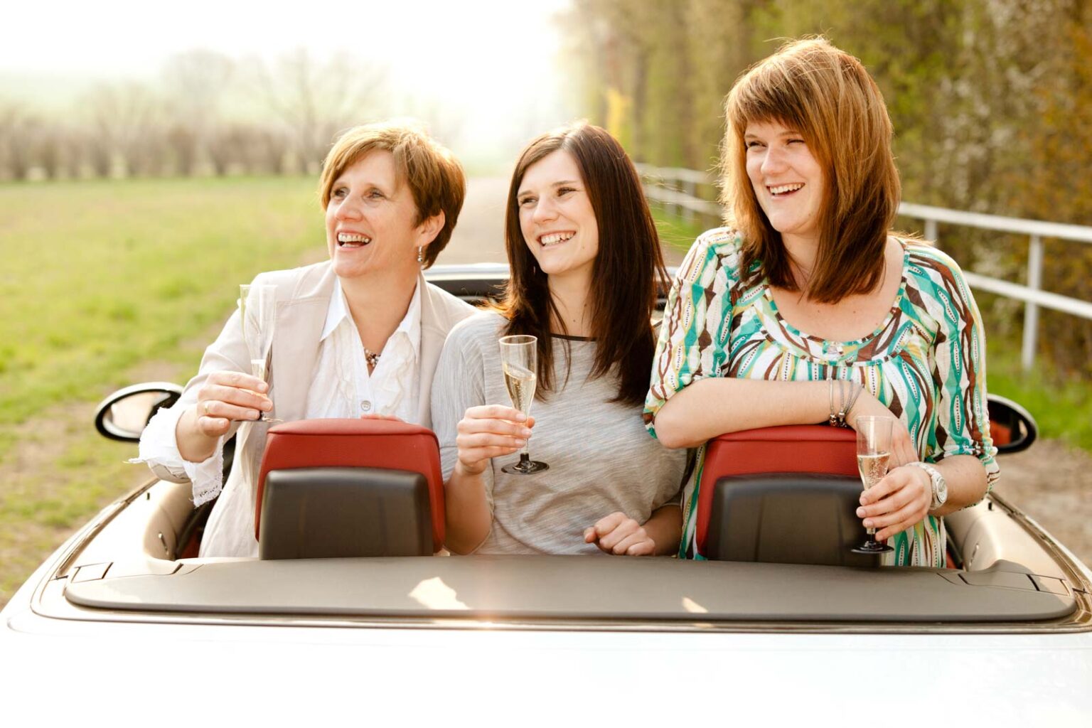Drei Frauen lachen und halten Champagnergläser in einem Cabrio, das in einer ländlichen Gegend auf einem Feldweg steht.