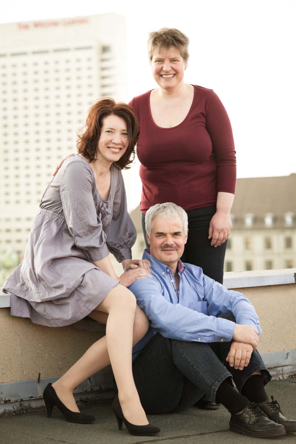 Drei Personen posieren auf einem Dach; zwei Frauen stehen und knien, während ein Mann davor sitzt. Im Hintergrund ist ein hohes Gebäude zu sehen.