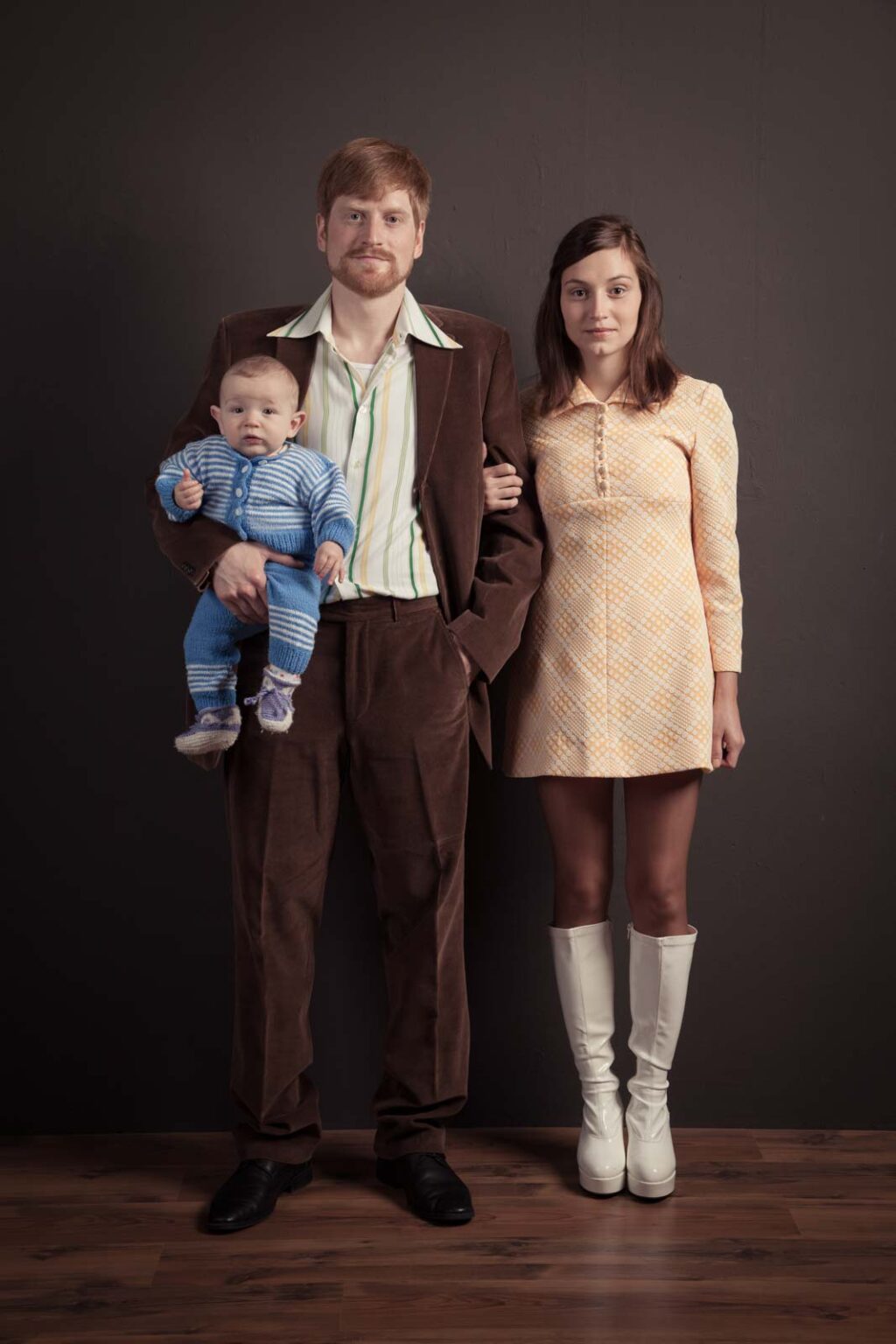 Ein Mann in einem braunen Anzug hält ein Baby in einem blauen Strampler, während eine Frau in einem gelben Kleid mit weißen Stiefeln neben ihm steht. Alle drei Personen posieren vor einem neutralen Hintergrund.