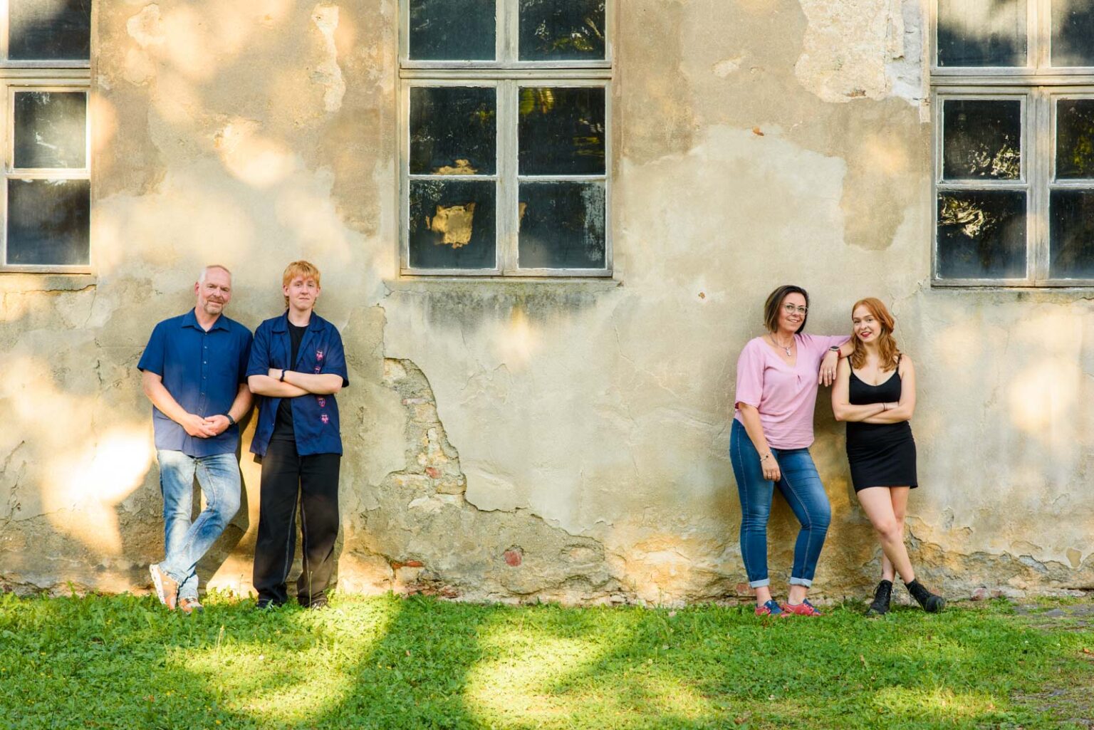 Vier Personen stehen an einer alten, verputzten Hauswand mit Fenstern; zwei Personen auf der linken Seite tragen blaue Hemden und Jeans, während die zwei Personen auf der rechten Seite in einem rosa Oberteil und einem schwarzen Kleid posieren. Sie stehen auf grasbewachsenem Boden im Schatten.