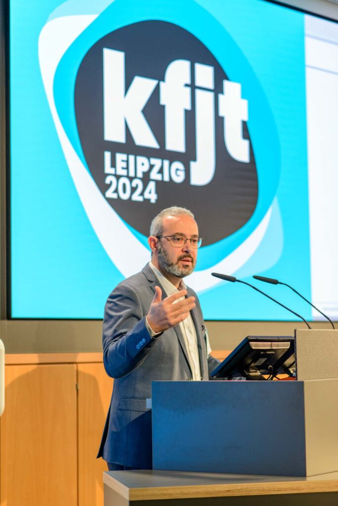 Ein Mann in einem blauen Anzug hält eine Präsentation an einem Rednerpult, auf dem Bildschirm hinter ihm steht "kfj Leipzig 2024".