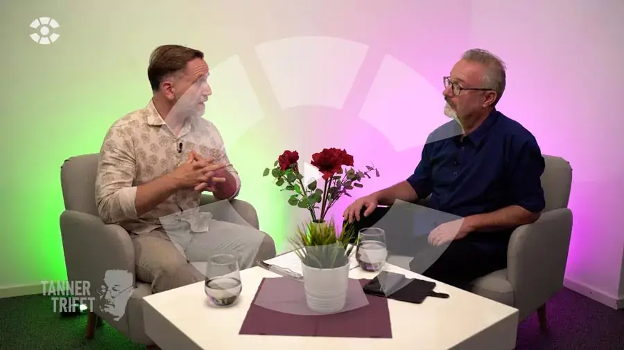 Zwei Männer im Gespräch, sitzen auf grauen Sesseln gegenüber an einem weißen Tisch mit roter Blumen- und grüner Topfpflanze, farbig hinterleuchtete Wand in Grün und Rosa, Casual-Kleidung, linke Person gestikuliert mit verschränkten Händen, rechte Person hört aufmerksam zu.