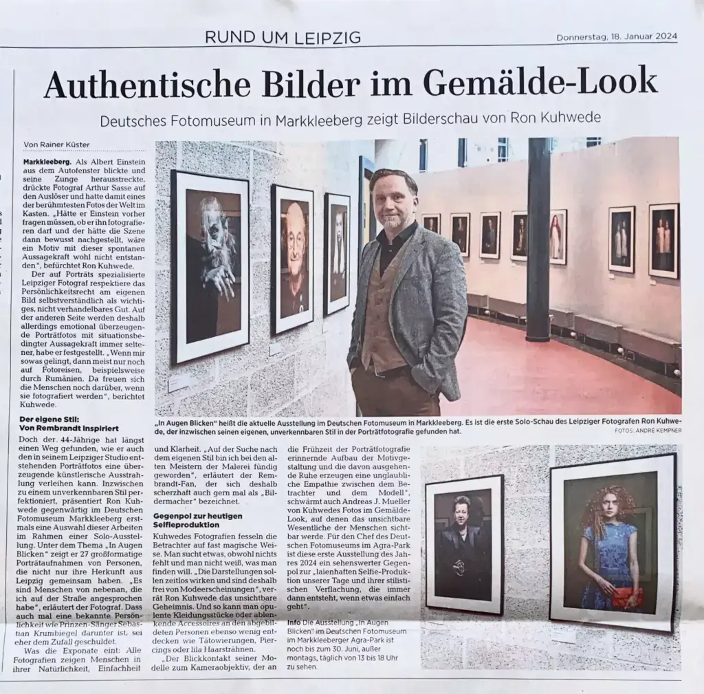 Zeitungsartikel mit Überschrift über eine Fotoausstellung von Ron Kuhwede im Deutschen Fotomuseum Markkleeberg, darunter ein Foto von Ron Kuhwede in der Ausstellung mit Porträtfotos an der Wand und weiteren Porträtfotos in Bilderrahmen daneben an einer weißen Wand.