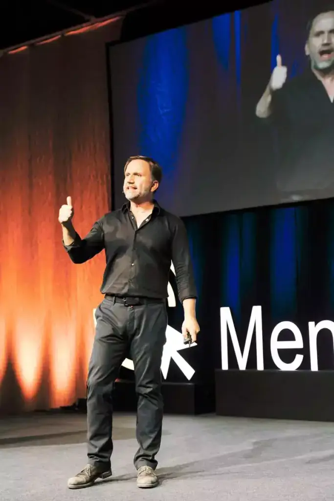 Mann in schwarzem Hemd und Hose steht auf einer Bühne, gibt Daumen hoch, vor einer leuchtend orange-blauen Wand mit einem großen Bildschirm und teilweisem Schriftzug im Hintergrund.
