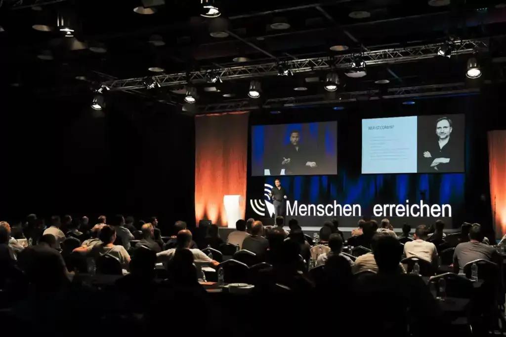 Ein Redner auf einer Bühne vor einem Publikum in einem dunklen Saal mit zwei großen Bildschirmen, einer zeigt den Sprecher, der andere eine Präsentationsfolie mit dem Titel "Wer ist Corwin?", und der Schriftzug "Menschen erreichen" leuchtet auf der Bühne.