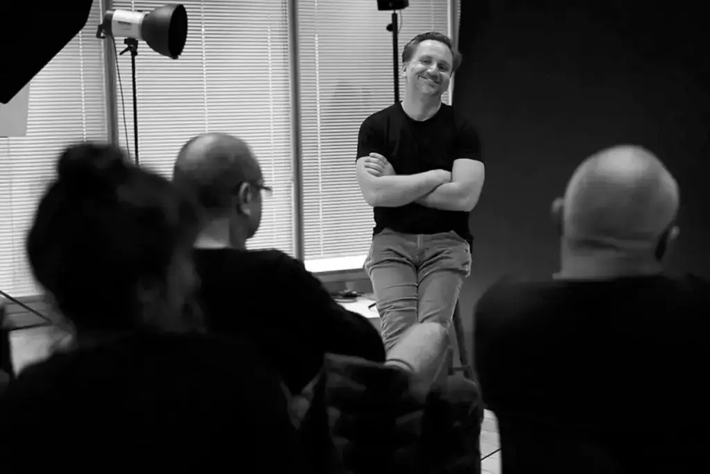 Schwarz-weiß-Foto einer sitzenden Gruppe von Personen, eine Person sitzt auf einem Stuhl mit verschränkten Armen und lächelt, im Hintergrund Fenster mit Jalousien und Fotostudio-Beleuchtung zu sehen.