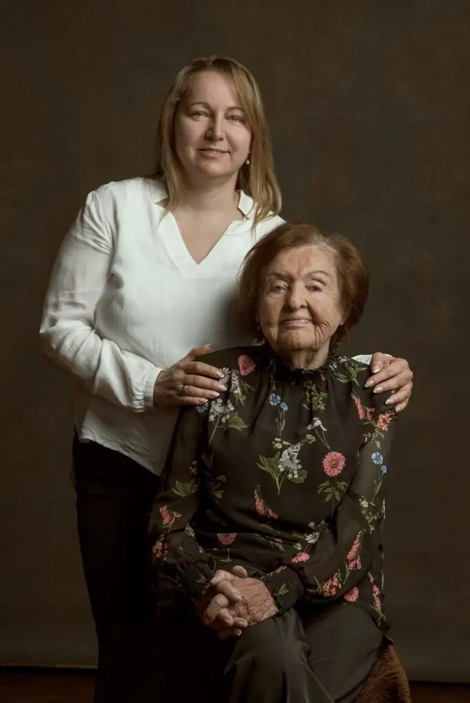 Porträt einer älteren Frau mit dunkler, geblümter Bluse, die sitzt, während eine jüngere Frau in weißer Bluse hinter ihr steht und ihre Schultern berührt, vor einfarbigem braunem Hintergrund.