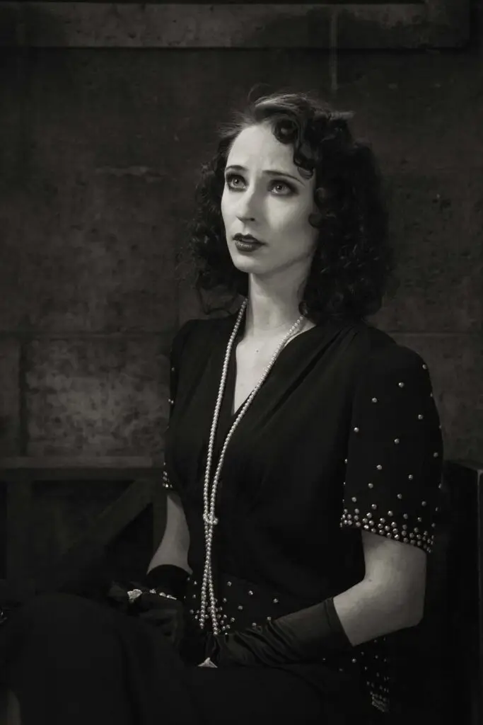 Schwarz-weiß-Porträt einer Frau mit lockigem Haar im Vintage-Stil, die ein schwarzes Kleid mit Perlenbesatz und lange schwarze Handschuhe trägt, blickt nach oben vor einem dunklen Hintergrund.