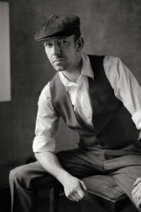 Schwarz-weiß-Porträt eines Mannes in Vintage-Kleidung mit Schiebermütze, Hemd mit hochgekrempelten Ärmeln und Weste, der auf einem Stuhl sitzt und direkt in die Kamera blickt.