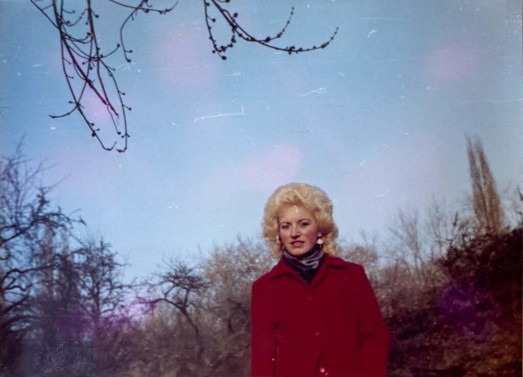 Frau mit blonden Locken und rotem Mantel vor einem winterlichen Hintergrund mit kahlen Bäumen und blauem Himmel