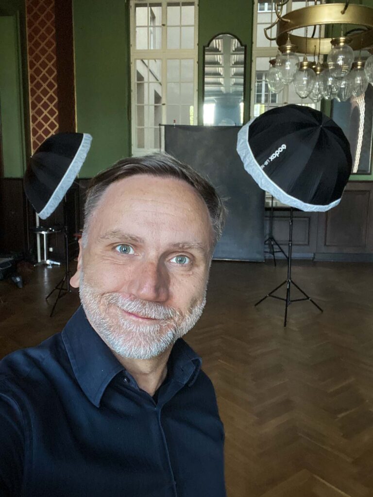 Mann mit grauem Bart und blauem Hemd macht ein Selfie in einem Raum mit hölzernem Parkettboden, zwei Fotolichtsoftboxen und einem großen Spiegel an der Wand im Hintergrund.