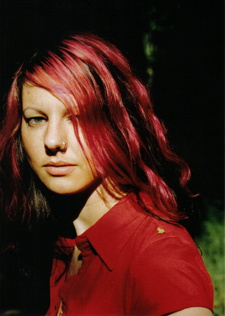 Porträt einer Frau mit langen, roten Haaren, die eine rote Bluse trägt, mit einem Nasenring und einem kleinen Schmetterling auf ihrer rechten Schulter, vor dunklem Hintergrund mit etwas grünem Licht im Hintergrund.