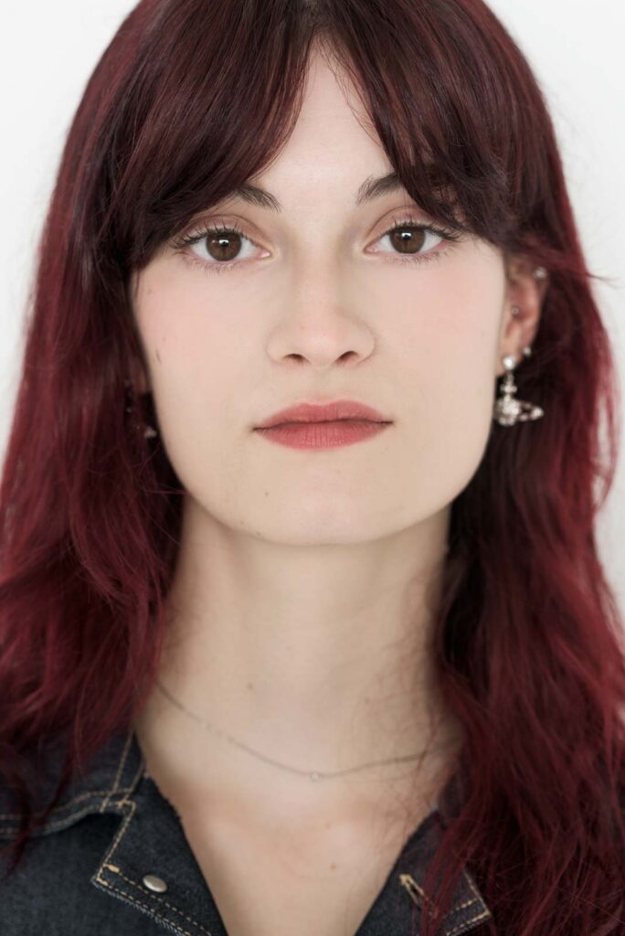 Porträt einer jungen Frau mit langen dunkelroten Haaren, braunen Augen, dezentem Make-up, silbernen Ohrringen und einem dunklen Denim-Oberteil vor weißem Hintergrund.