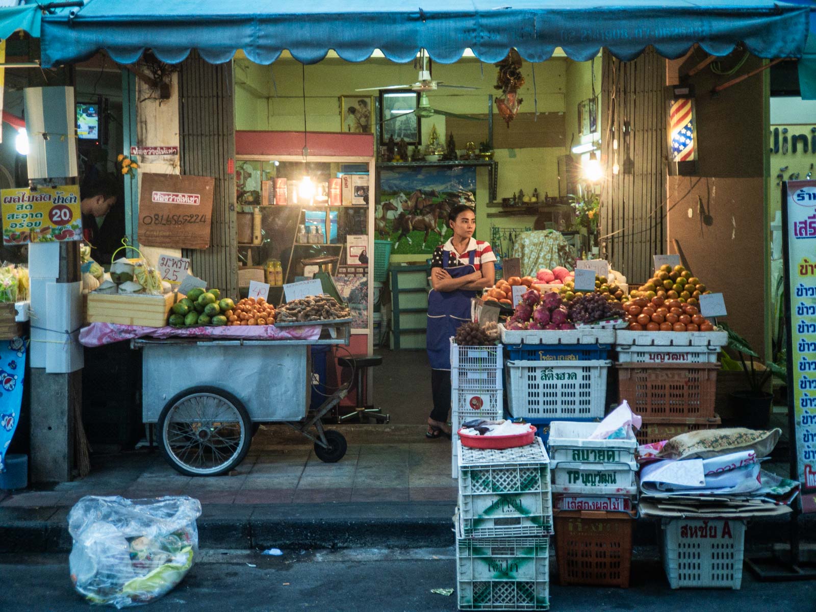 Straßenmarktstand mit einer Frau in einer Schürze, die vor einer Auswahl an frischem Obst und Gemüse steht, mit beleuchtetem Verkaufsraum im Hintergrund und blauer Markisenüberdachung.