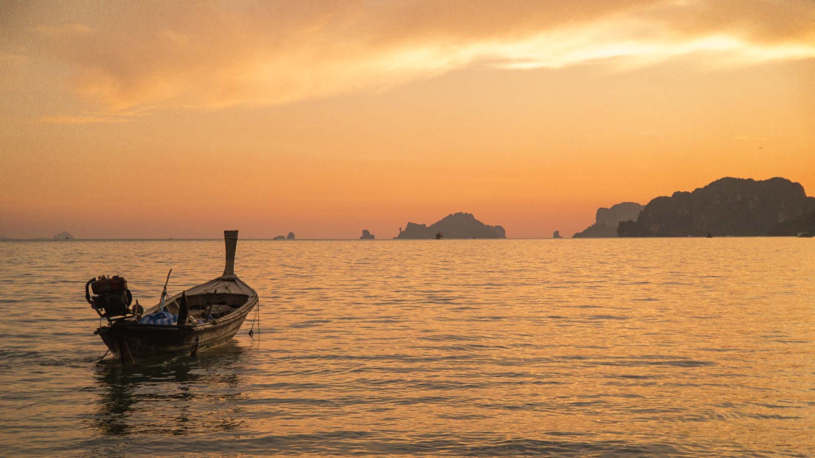 Ein traditionelles Holzboot auf ruhigem Wasser bei Sonnenuntergang mit orangefarbenem Himmel und einer Silhouette von Inseln am Horizont.