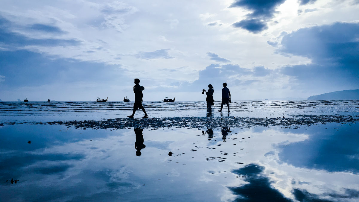 Silhouetten von vier Menschen, die am Ufer eines ruhigen Meeres bei Flut gehen, mit mehreren kleinen Booten im Hintergrund und bewölktem Himmel, der sich im Wasser spiegelt