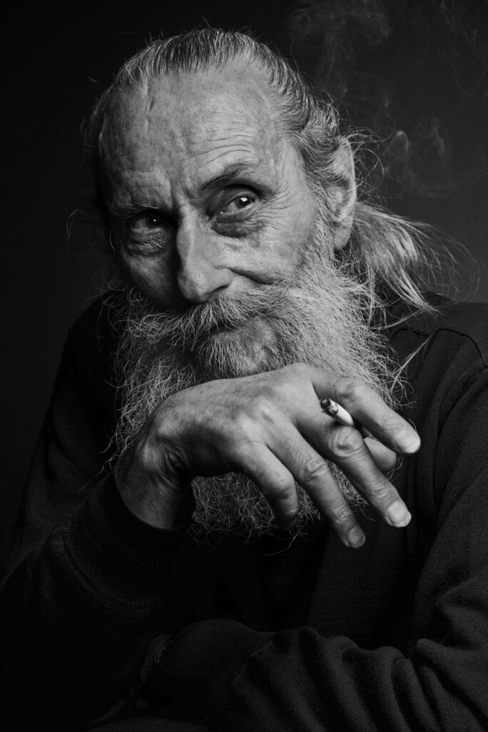 Schwarz-weiß-Porträt eines älteren Mannes mit langem, grauem Bart und gebundenem Haar, der nachdenklich nach rechts schaut und eine brennende Zigarette in der rechten Hand hält.