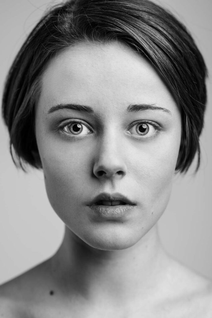 Schwarz-weiß-Porträt eines jungen Frauengesichts mit kurzem Haar, die direkt in die Kamera schaut, mit offenem Mund und neutralem Ausdruck vor schlichtem Hintergrund.