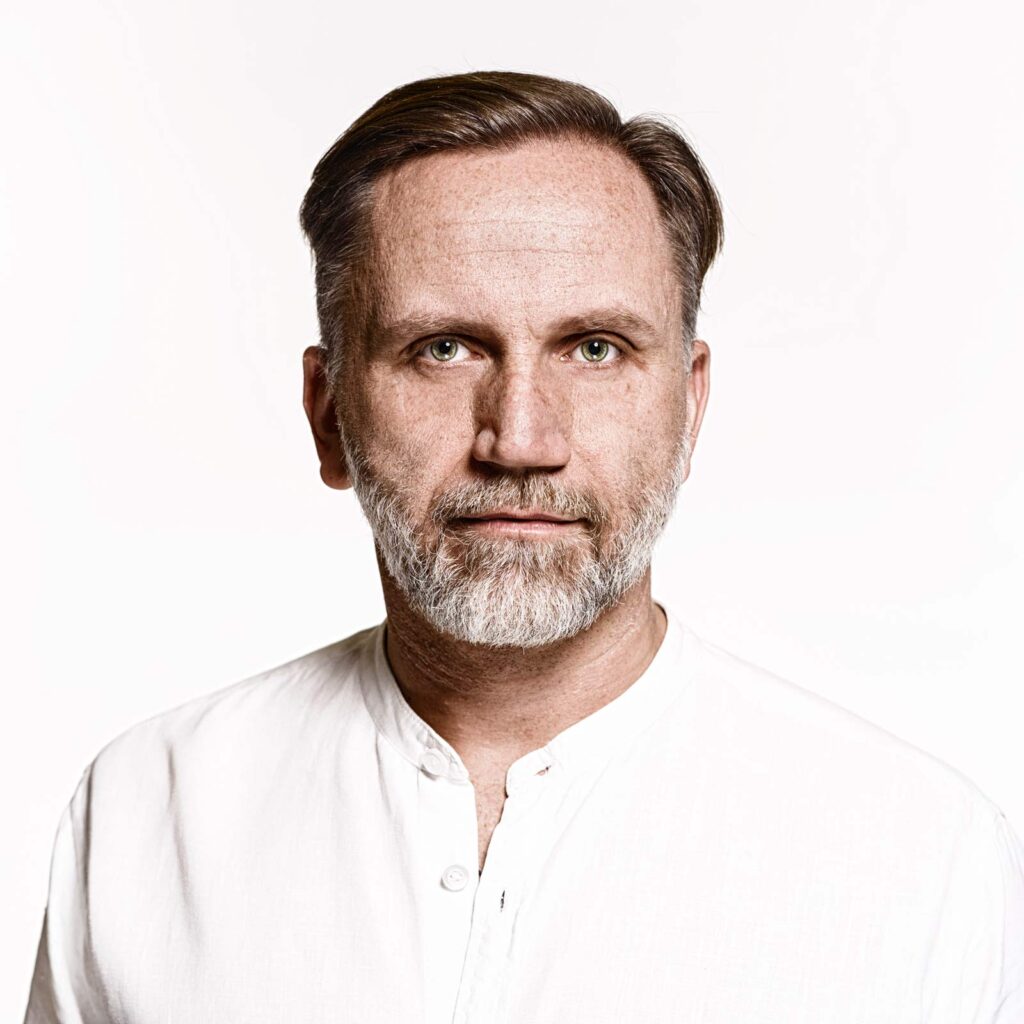 Porträt eines mittelalten Mannes mit grauem Bart und zurückgekämmtem braunem Haar, der ein weißes Hemd mit Stehkragen trägt und direkt in die Kamera schaut, vor weißem Hintergrund.