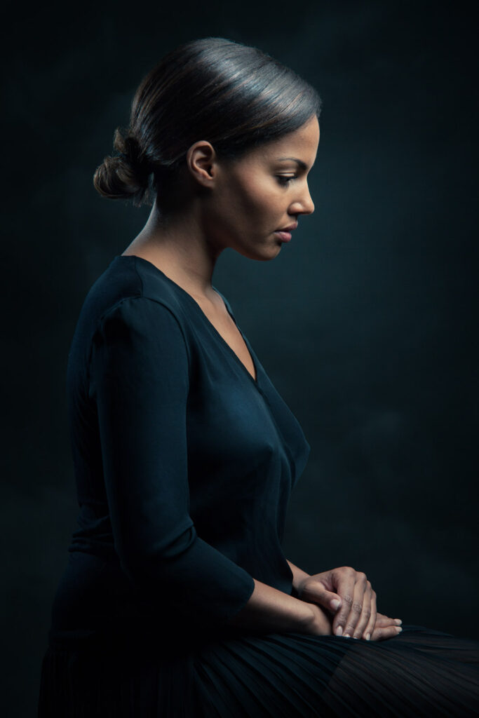Porträt einer Frau in Seitenansicht vor schwarzem Hintergrund, die nachdenklich mit gefalteten Händen sitzt und eine schwarze Bluse trägt.