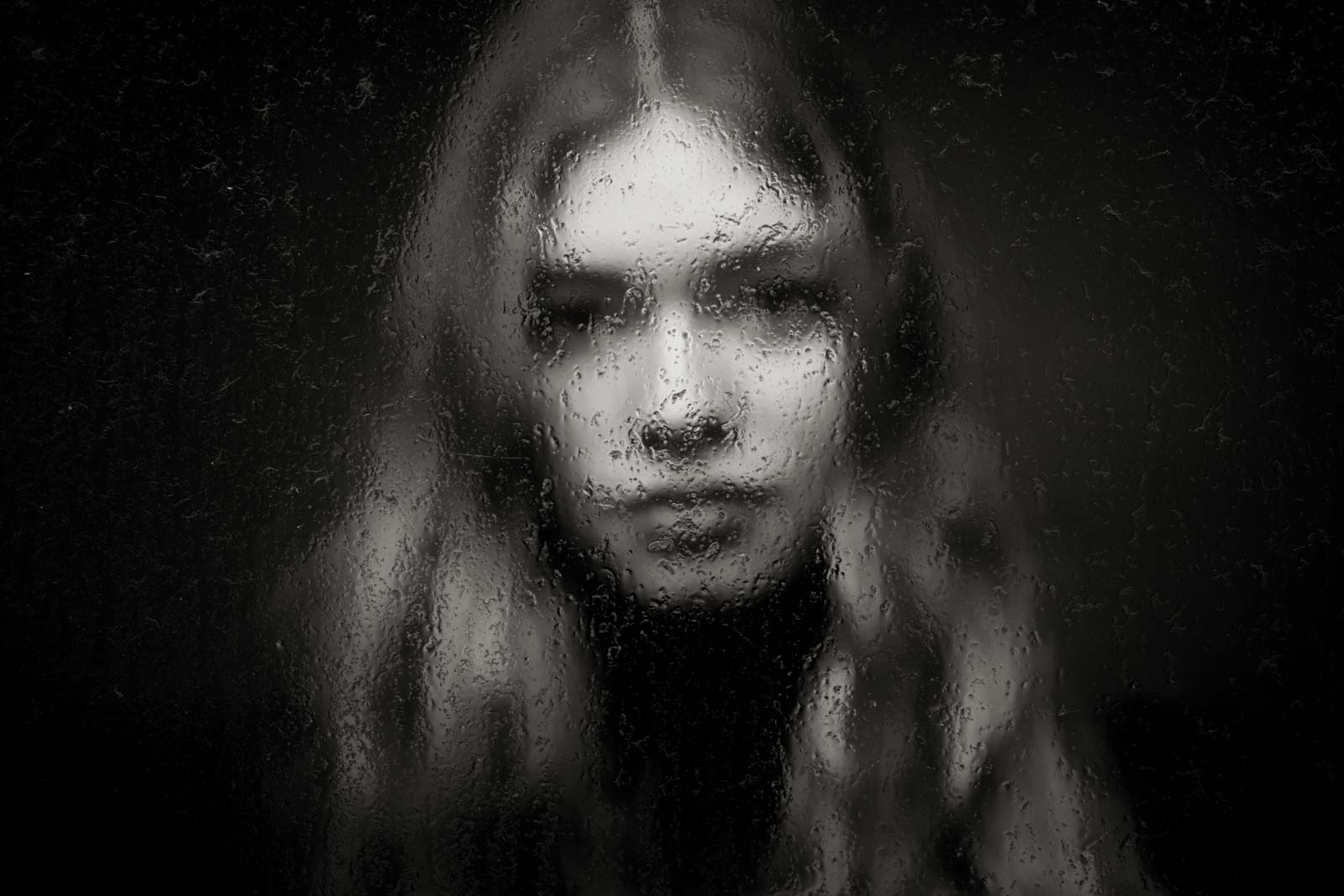Portrait einer Frau mit langen Haaren hinter einer regennassen Glasscheibe in Schwarz-Weiß, die ihr Gesicht verschwommen zeigt.