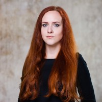 Frau mit langem roten Haar und schwarzem Oberteil vor unscharfem, hellem Hintergrund.