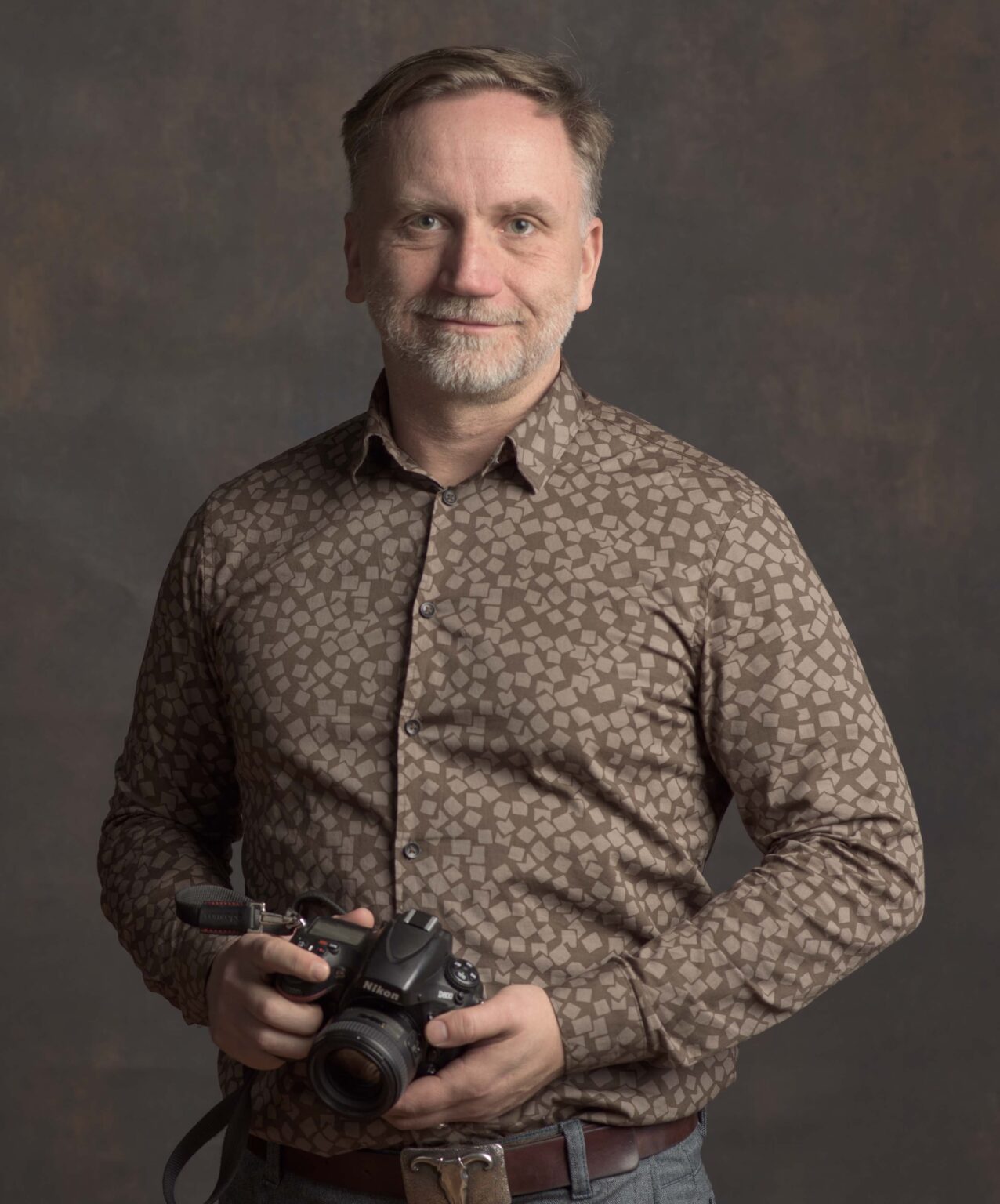 Mann mit grauem Bart und gemustertem Hemd hält eine digitale Spiegelreflexkamera in den Händen.